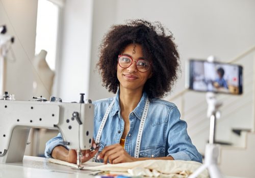 Pretty African-American woman blogger with scissors and glasses at workplace in sewing workshop